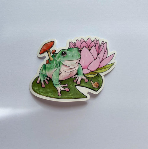 A die-cut sticker featuring an illustrated green frog sitting on a lily pad next to a pink water lily and a small orange mushroom, shown on a plain light background.